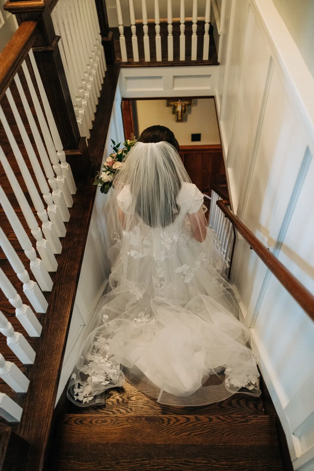 Viewed from above, the bride descends a wooden staircase with her veil and train flowing behind her, a crucifix visible on the wall below. — Tim Larsen Photography, Brainerd Lakes MN