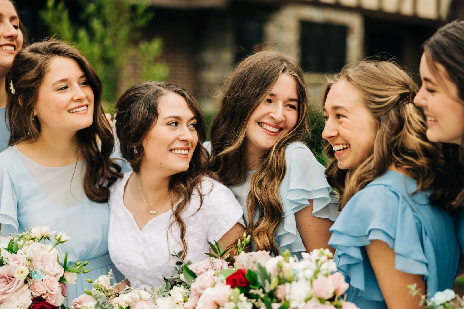 The bride laughs with five bridesmaids in dusty-blue dresses gathered closely around her, all holding pastel bouquets outside a stone house. — Tim Larsen Photography, Brainerd Lakes MN