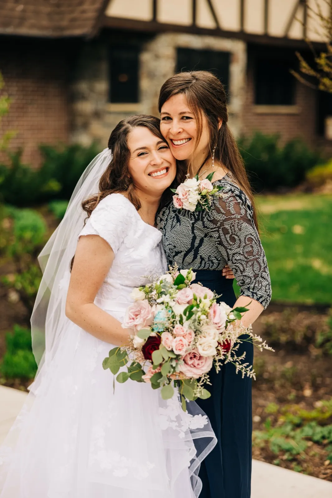 The bride and a female family member press cheeks together and smile broadly while the bride holds a large pink and white bouquet in front of a Tudor-style stone building. — Tim Larsen Photography, Brainerd Lakes MN