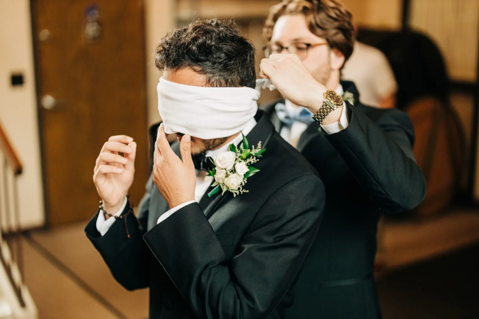 A groomsman ties a white blindfold around the groom's eyes as the groom stands in his tuxedo with a white rose boutonniere. — Tim Larsen Photography, Brainerd Lakes MN