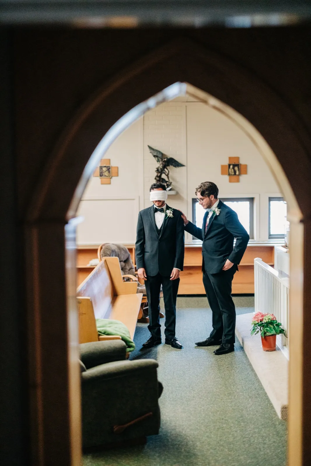 The blindfolded groom stands waiting with a groomsman's hand on his shoulder, viewed through a Gothic archway from an adjacent room. — Tim Larsen Photography, Brainerd Lakes MN