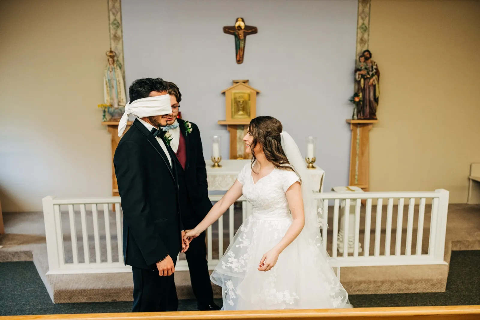 A blindfolded groom reaches for the bride's hand at a Catholic church altar beneath a crucifix as she smiles toward him in her white gown and veil. — Tim Larsen Photography, Brainerd Lakes MN