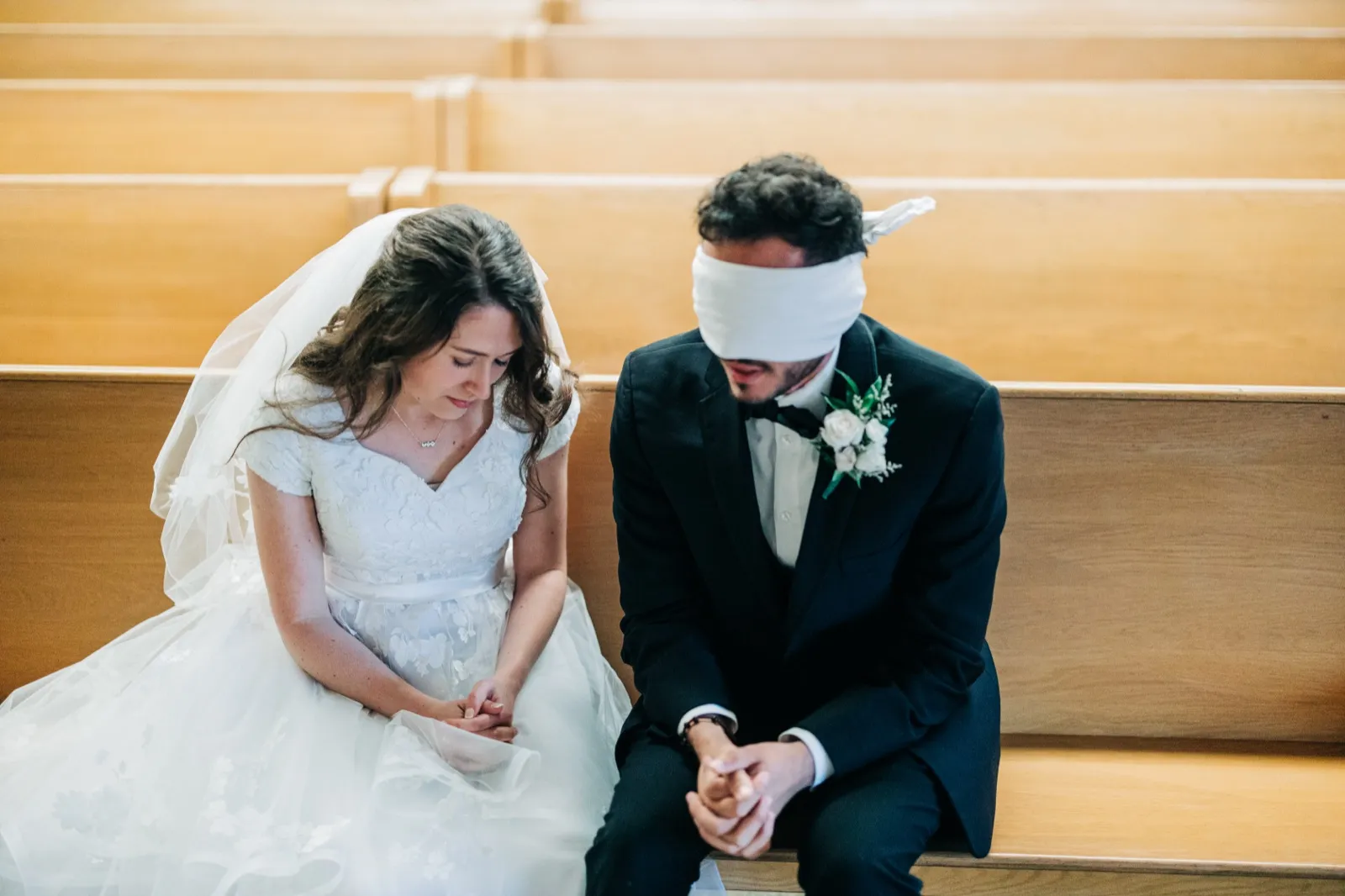 From a high angle, the bride and blindfolded groom sit side by side in a wooden church pew with heads bowed and hands clasped in prayer. — Tim Larsen Photography, Brainerd Lakes MN