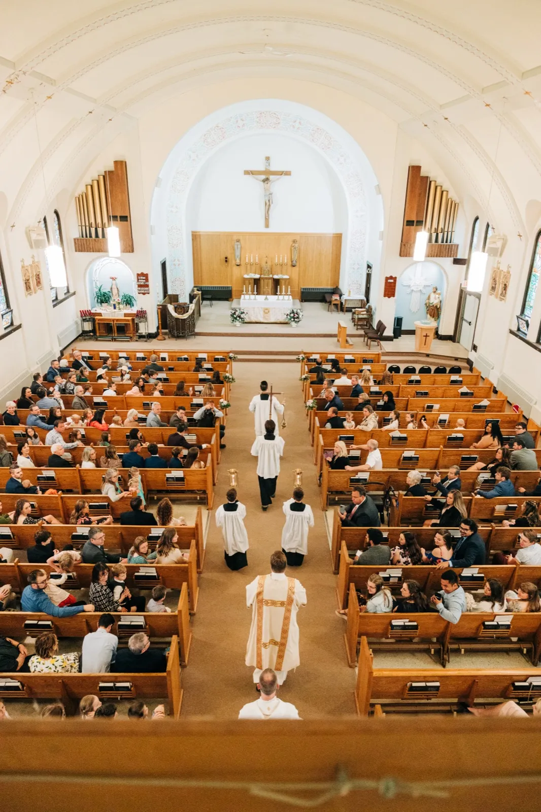An elevated balcony view down the center aisle of a Catholic church as a priest, altar servers, and a crucifer process toward the altar beneath a large crucifix, with guests filling the pews on either side. — Tim Larsen Photography, Brainerd Lakes MN