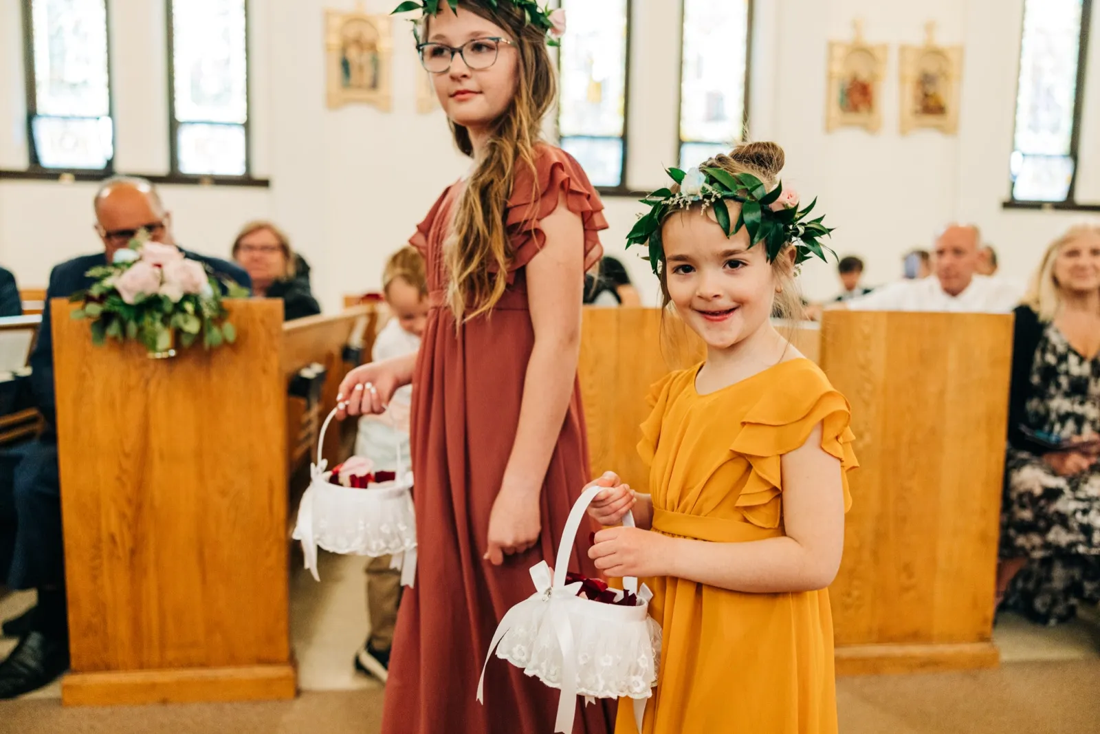 Two flower girls in floral crowns hold lace baskets in a church aisle, the younger one in a yellow dress smiling at the camera while the older one in a rust-colored dress looks past it. — Tim Larsen Photography, Brainerd Lakes MN