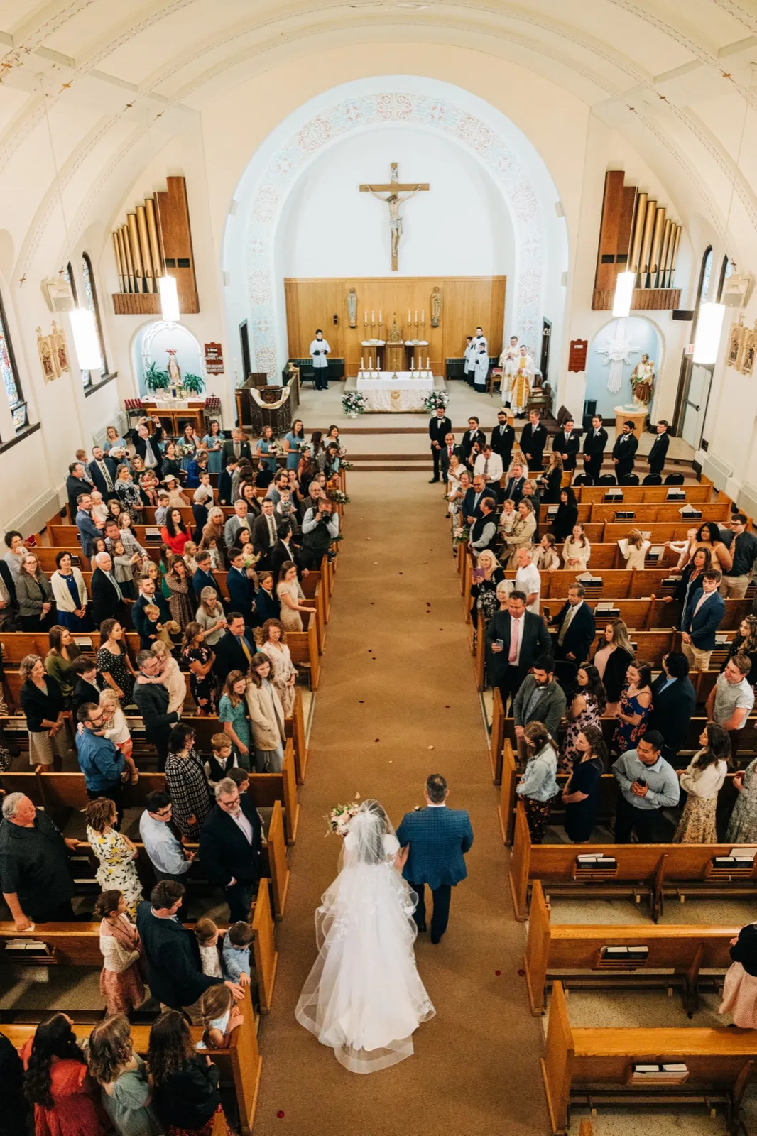 An elevated wide view down the aisle of an ornate Catholic church shows the bride and groom walking together toward the camera, her veil trailing behind, with full pews of guests on either side and a crucifix above the altar in the distance. — Tim Larsen Photography, Brainerd Lakes MN
