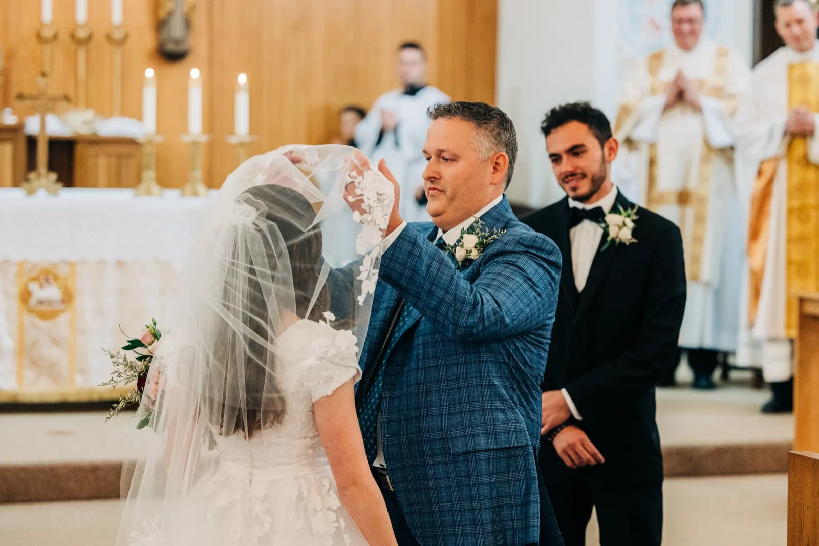 The groom lifts the bride's lace-edged veil at a church altar as the priest and a groomsman stand reverently behind them. — Tim Larsen Photography, Brainerd Lakes MN