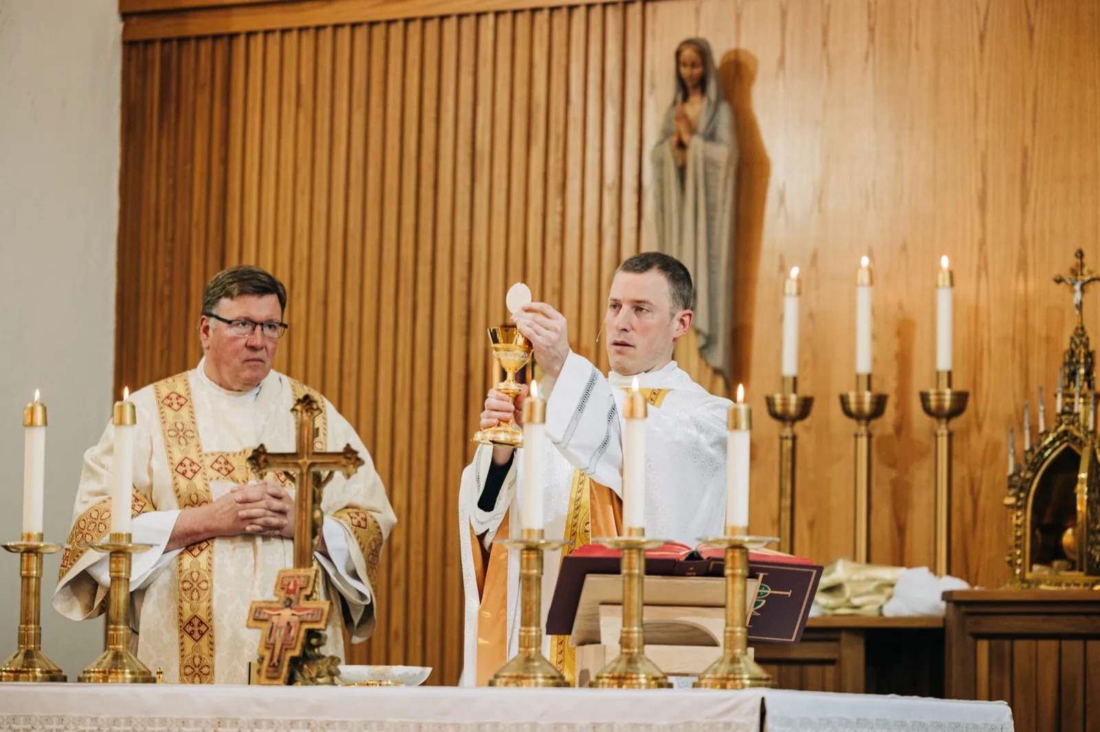A priest elevates the consecrated host above a golden chalice during a Catholic wedding mass, with a second clergy member standing beside him and a statue of the Virgin Mary on the wood-paneled wall behind. — Tim Larsen Photography, Brainerd Lakes MN