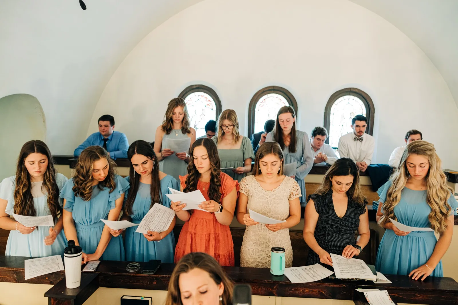 A group of bridesmaids in dusty blue dresses and groomsmen in shirts and ties stand singing from sheet music beneath an arched chapel ceiling and stained-glass windows. — Tim Larsen Photography, Brainerd Lakes MN