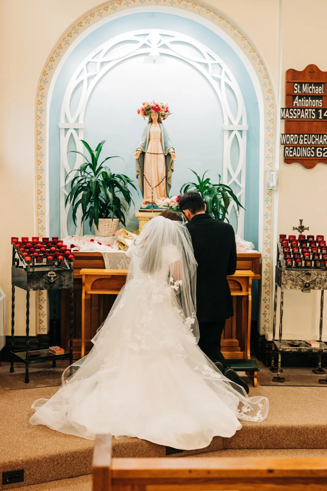 The bride and groom kneel together before a statue of Mary at a side altar in St. Joseph's church, the bride's veil and lace train spreading across the carpeted steps. — Tim Larsen Photography, Brainerd Lakes MN