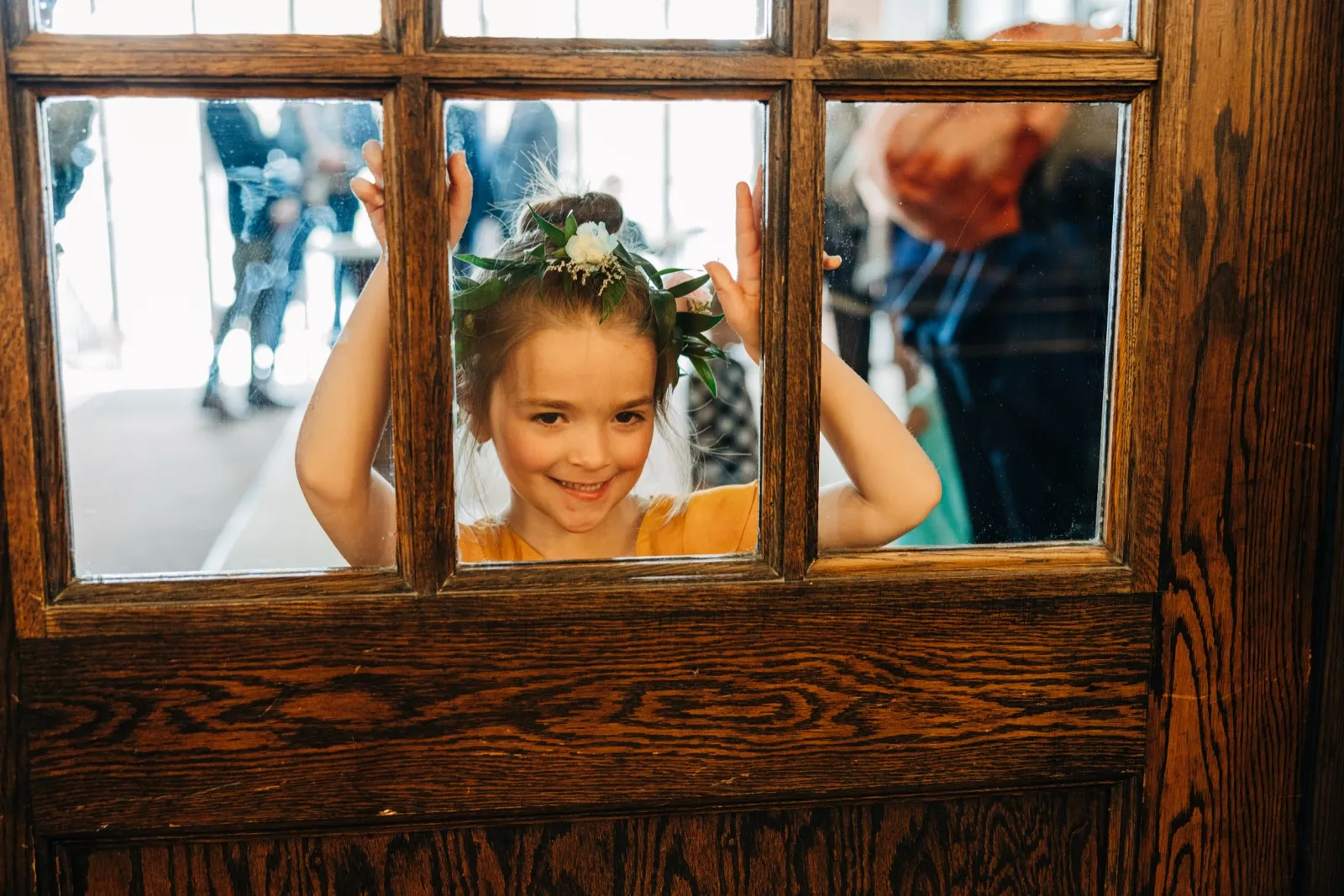 A flower girl wearing a floral crown peeks through the glass pane of a wooden door, hands pressed against the frame. — Tim Larsen Photography, Brainerd Lakes MN