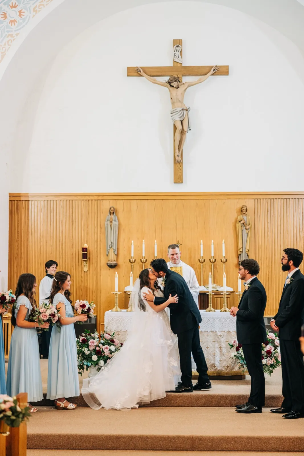 The bride and groom share their first kiss at the altar beneath a large crucifix, with the priest smiling behind them and the bridal party standing on either side. — Tim Larsen Photography, Brainerd Lakes MN