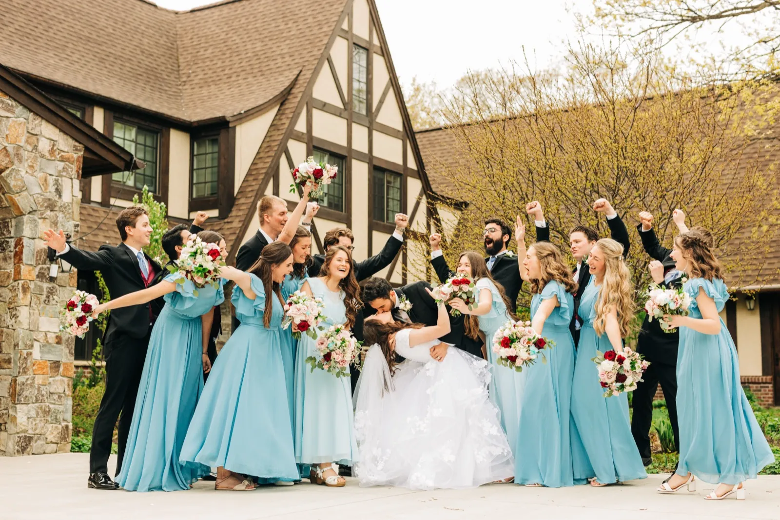 The bride and groom kiss at the center of their cheering bridal party, with bridesmaids in dusty-blue gowns and groomsmen in dark suits raising their arms in front of a Tudor-style lodge. — Tim Larsen Photography, Brainerd Lakes MN