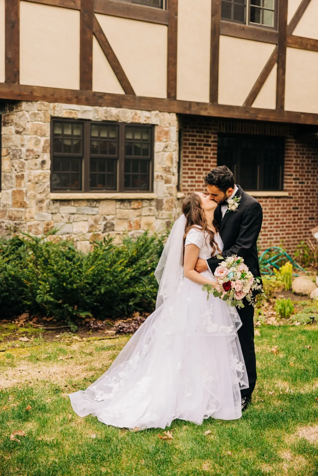 The bride and groom kiss on a lawn outside a Tudor-style building, her veil and dress train spreading across the grass while she holds a pink and white bouquet. — Tim Larsen Photography, Brainerd Lakes MN