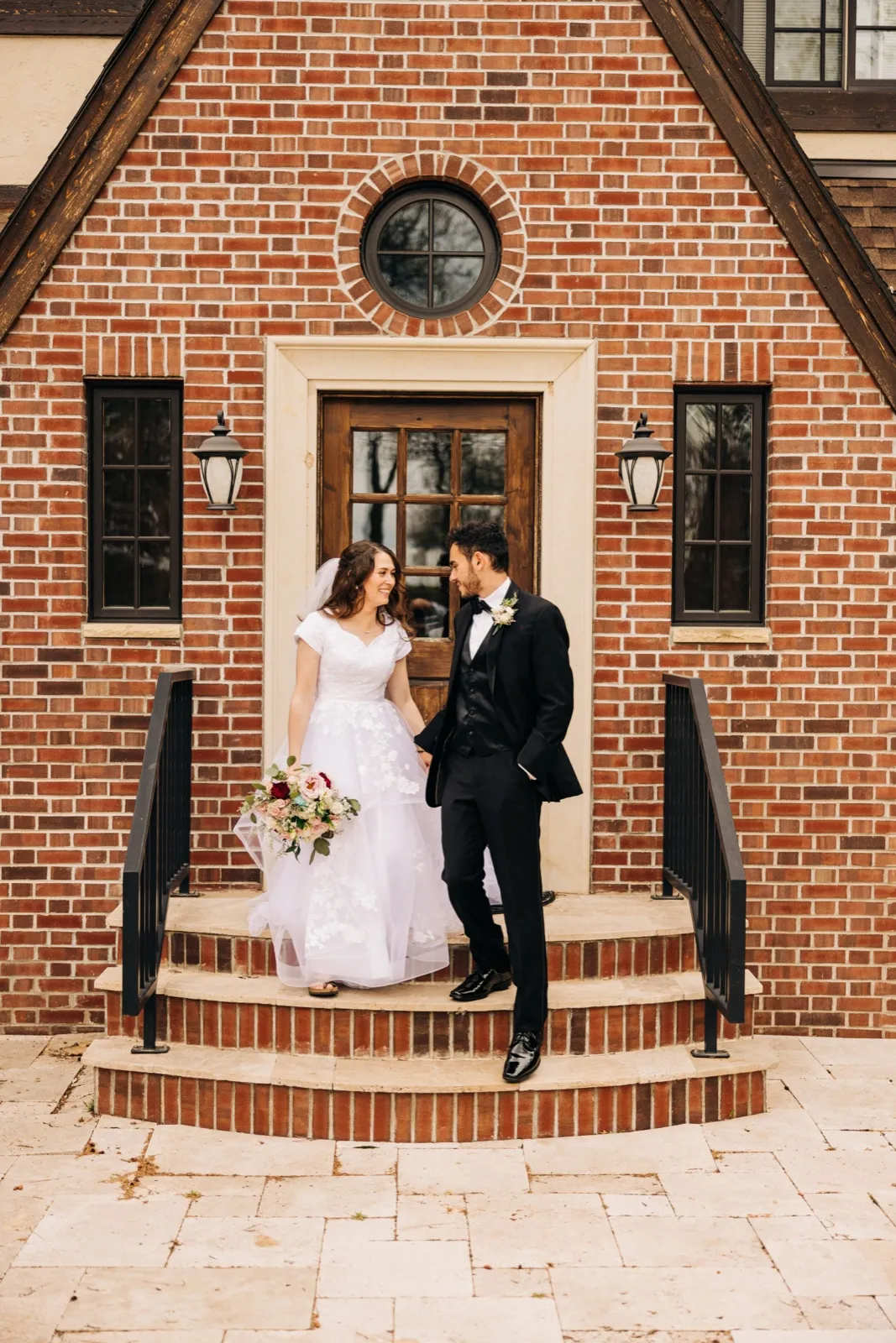 The bride and groom walk down brick front steps holding hands, smiling at each other beneath a Tudor-style brick facade with a round window above the doorway. — Tim Larsen Photography, Brainerd Lakes MN