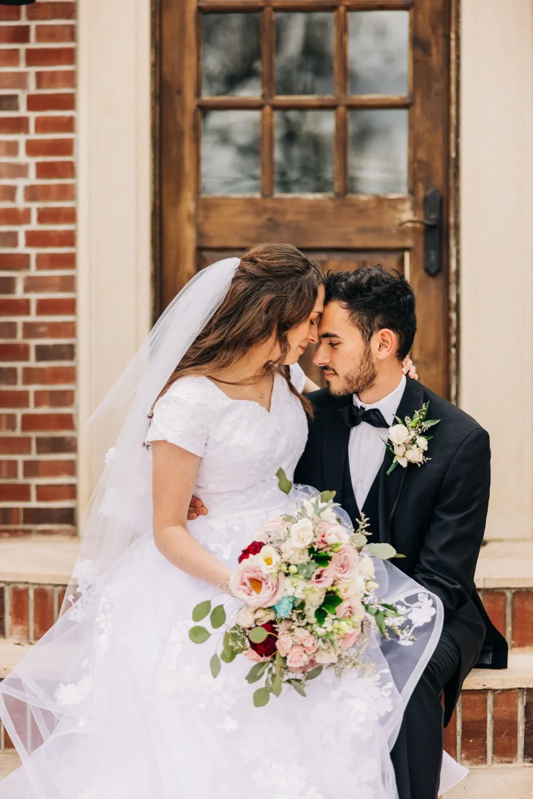 The bride and groom sit close on brick steps in front of a paneled wooden door, foreheads touching as she holds a large pink and red bouquet across her lap. — Tim Larsen Photography, Brainerd Lakes MN