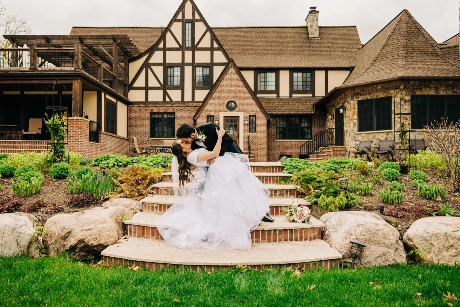 The groom dips the bride into a kiss on the stone steps of a Tudor-style lake home, her dress and veil spilling down the stairs into the surrounding gardens. — Tim Larsen Photography, Brainerd Lakes MN