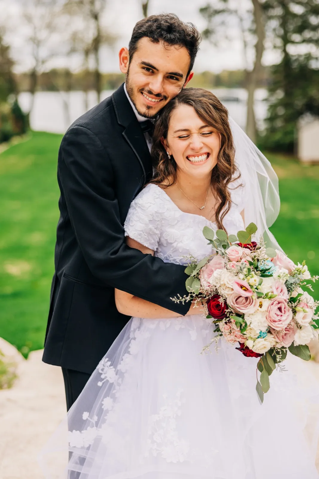 The groom embraces the bride from behind on a lakeside lawn as she squeezes her eyes shut in laughter, holding a large pink and white bouquet. — Tim Larsen Photography, Brainerd Lakes MN