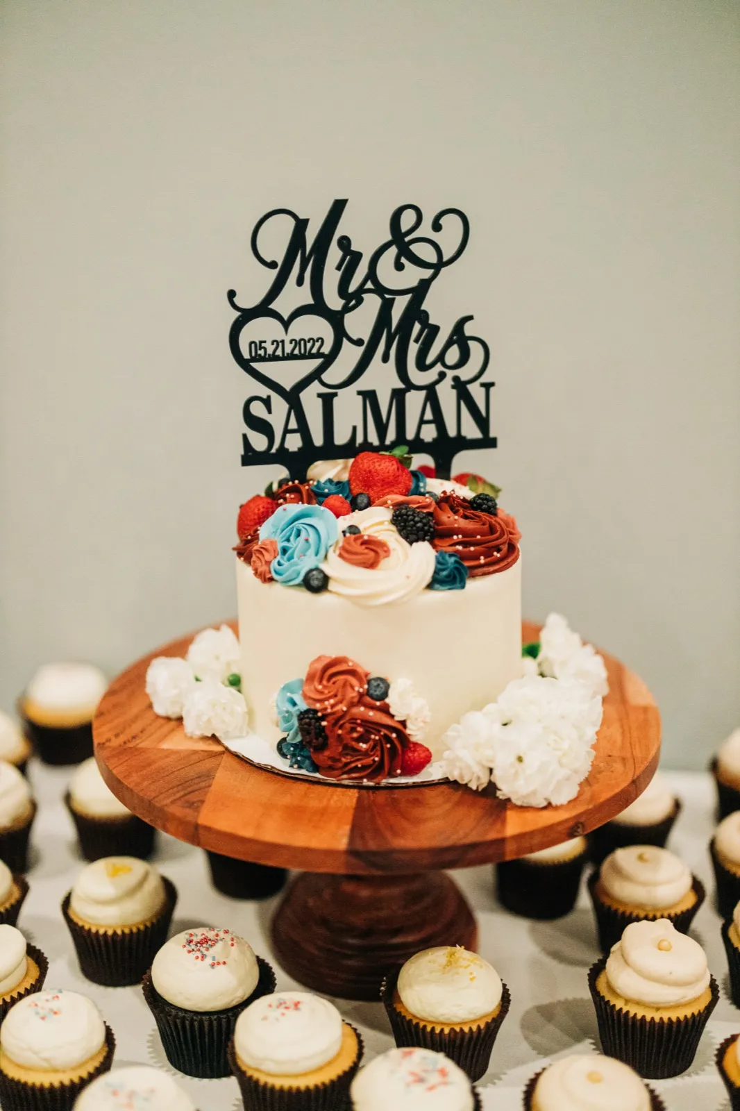 A small white wedding cake topped with red and blue buttercream flowers, berries, and a 'Mr & Mrs Salman 05.21.2022' cake topper, sits on a wooden cake stand surrounded by rows of cupcakes. — Tim Larsen Photography, Brainerd Lakes MN