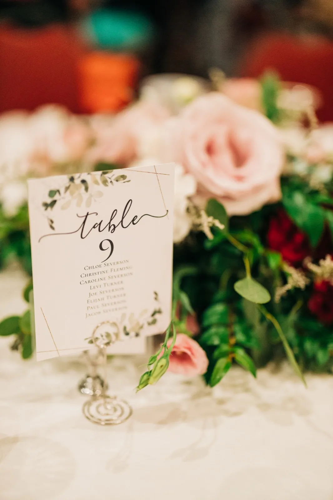 A calligraphed Table 9 seating card stands on a silver swirled holder in front of a centerpiece of blush roses, burgundy blooms, and trailing greenery. — Tim Larsen Photography, Brainerd Lakes MN