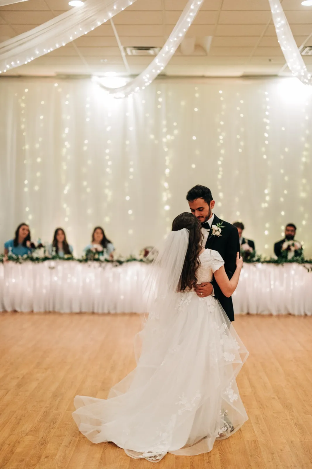 The bride and groom share their first dance on a wood floor, her veil and train trailing behind them, with the wedding party watching from a draped head table backed by twinkle lights. — Tim Larsen Photography, Brainerd Lakes MN