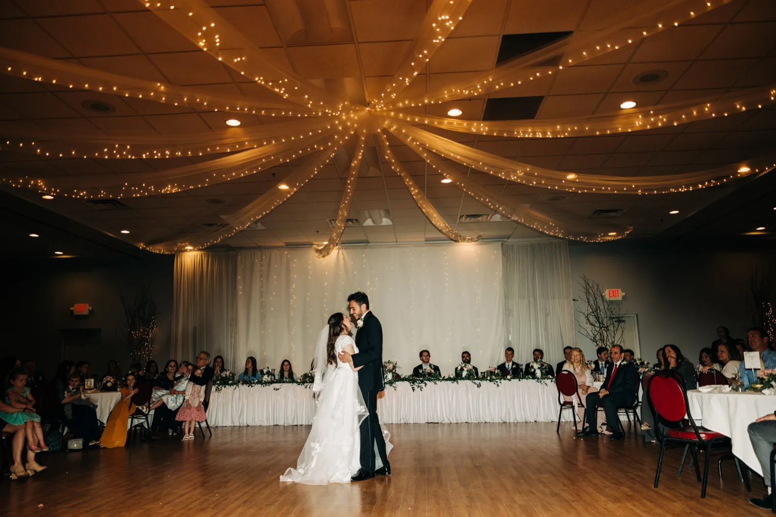The couple share their first dance in the center of a reception hall beneath a radiating canopy of string lights, with the bridal party and guests watching from a long head table behind them. — Tim Larsen Photography, Brainerd Lakes MN