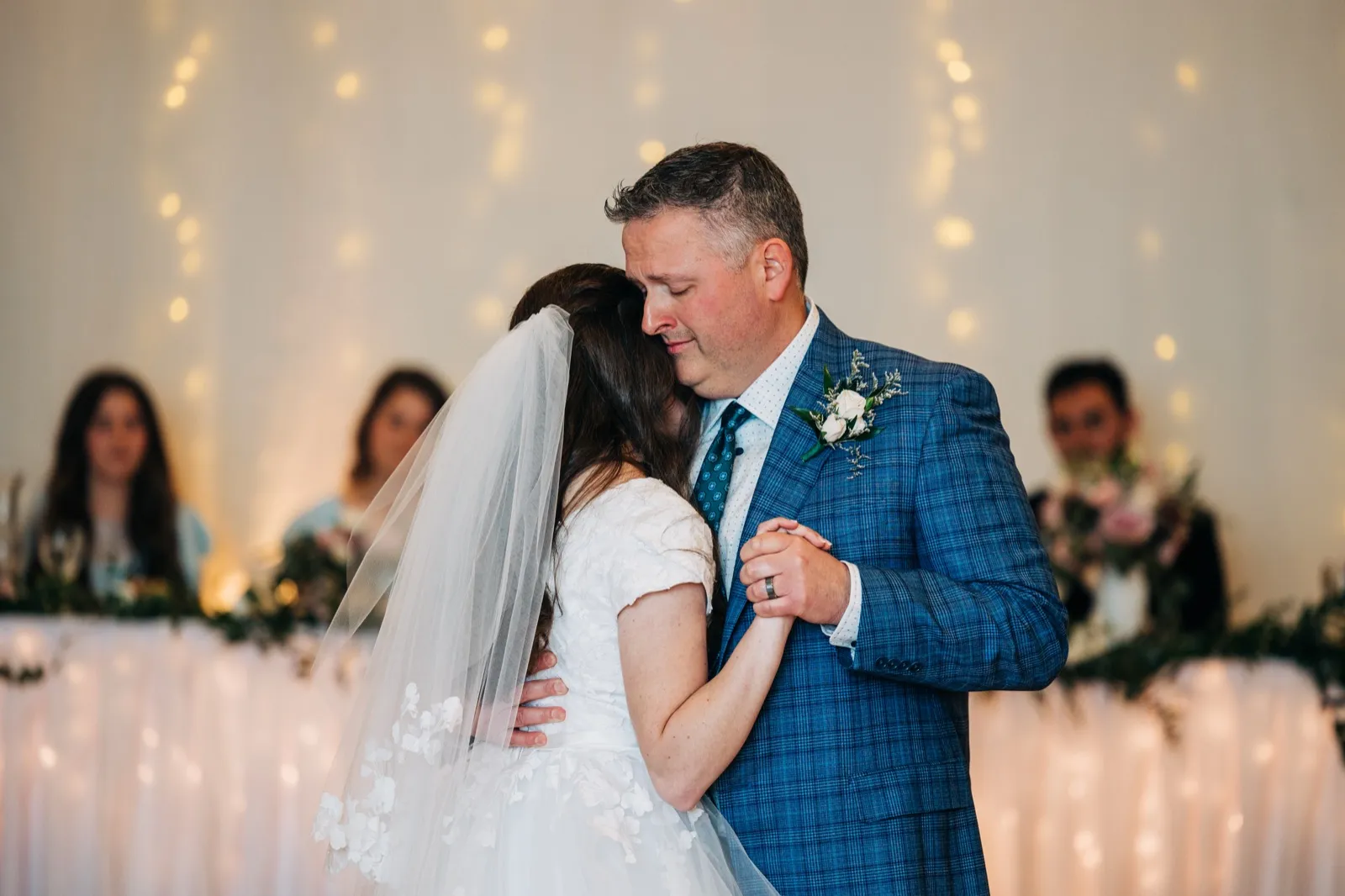 The groom rests his head against the bride during a slow dance, holding her hand as guests watch from the head table draped in string lights behind them. — Tim Larsen Photography, Brainerd Lakes MN