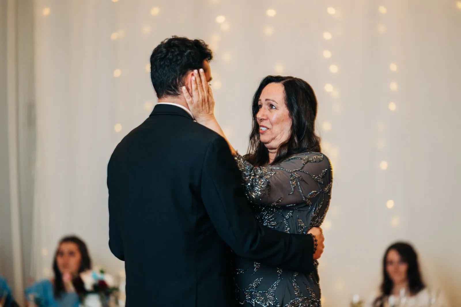 The groom's mother cradles his face in her hand during their dance, her eyes welling with emotion in front of a string-light backdrop. — Tim Larsen Photography, Brainerd Lakes MN
