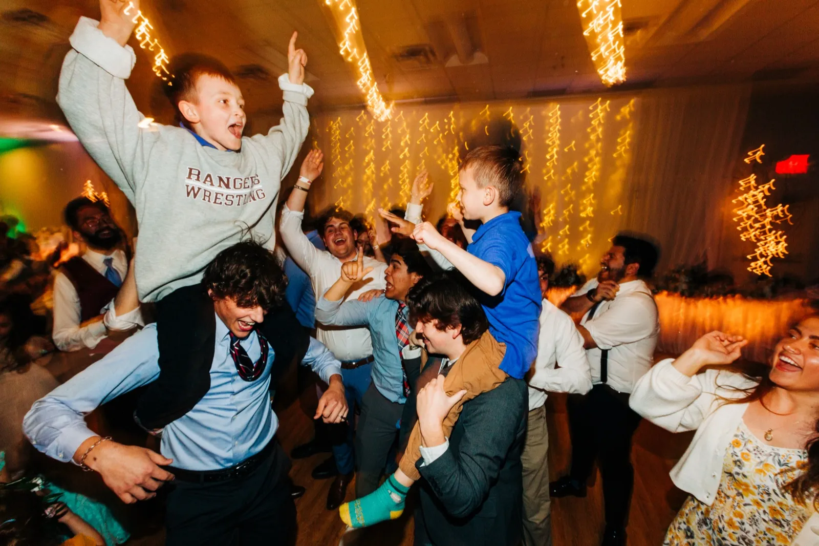 Two boys ride on the shoulders of older guests on a packed dance floor, arms raised and shouting, with string lights streaking behind them. — Tim Larsen Photography, Brainerd Lakes MN