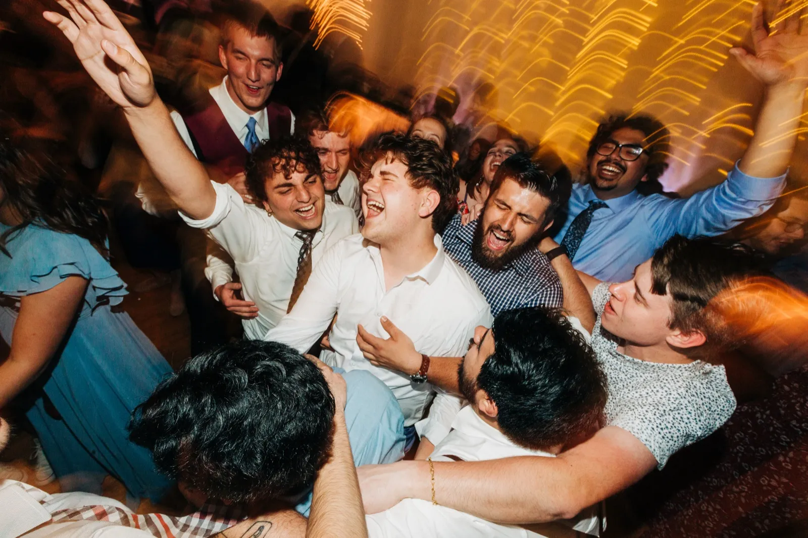 The groom is surrounded by friends belting out a song together on the dance floor as warm light streaks blur around them. — Tim Larsen Photography, Brainerd Lakes MN