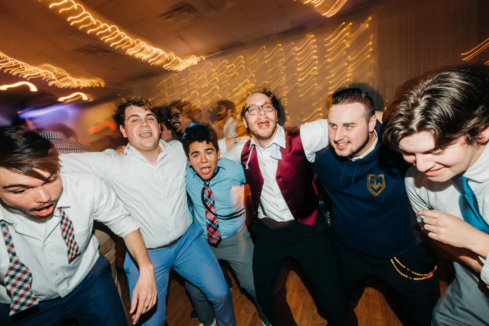 The groom and a tight huddle of groomsmen and guests sing into the camera with arms around each other on the dance floor, streaks of warm string lights blurred behind them. — Tim Larsen Photography, Brainerd Lakes MN