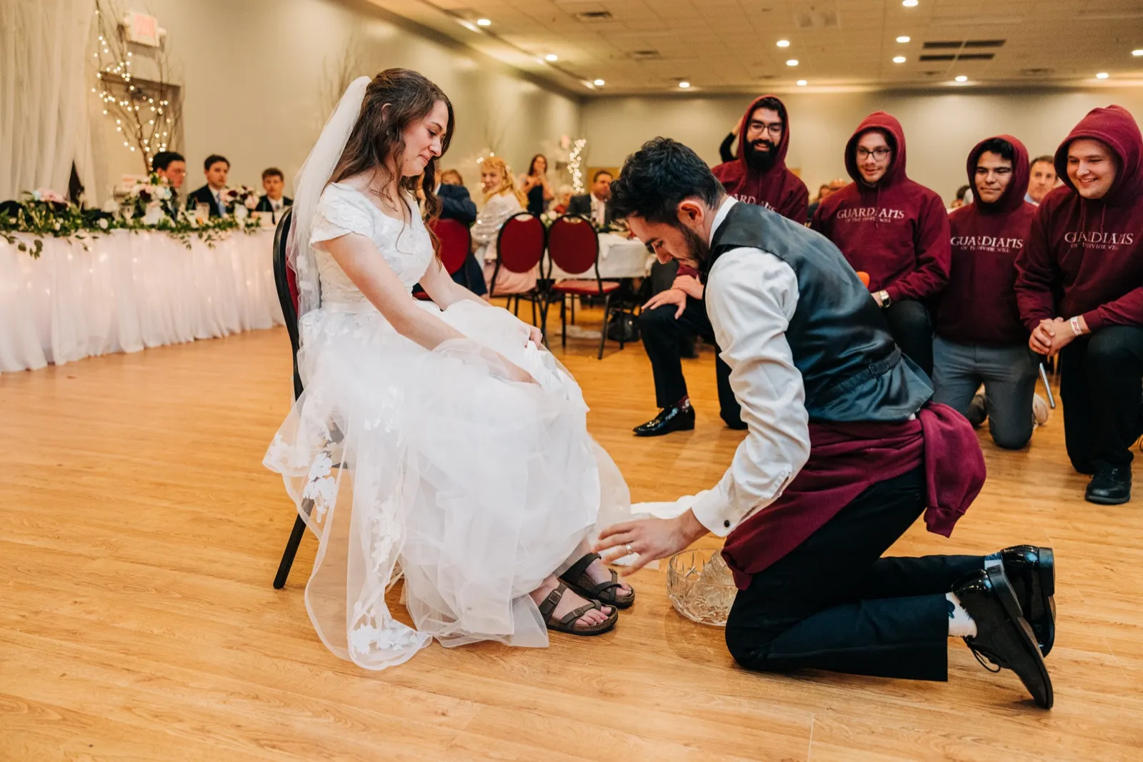 The groom kneels on the dance floor to wash the bride's feet from a glass bowl while a row of groomsmen in matching maroon hoodies kneel watching. — Tim Larsen Photography, Brainerd Lakes MN