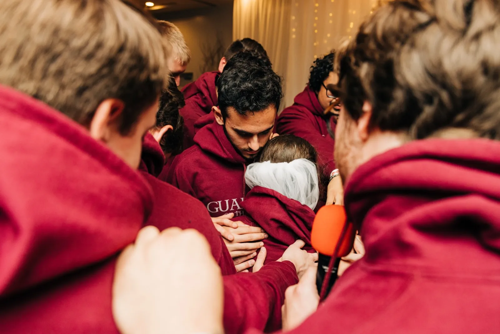 The groom embraces the bride at the center of a tight huddle of groomsmen in maroon hoodies, heads bowed in prayer. — Tim Larsen Photography, Brainerd Lakes MN