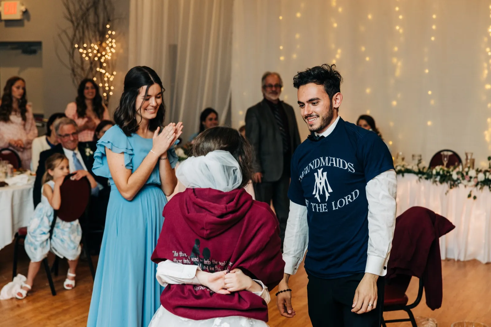 The bride pulls on a maroon hooded sweatshirt with religious lettering while the groom stands beside her in a matching navy 'Handmaids of the Lord' t-shirt, with a bridesmaid in blue laughing nearby. — Tim Larsen Photography, Brainerd Lakes MN