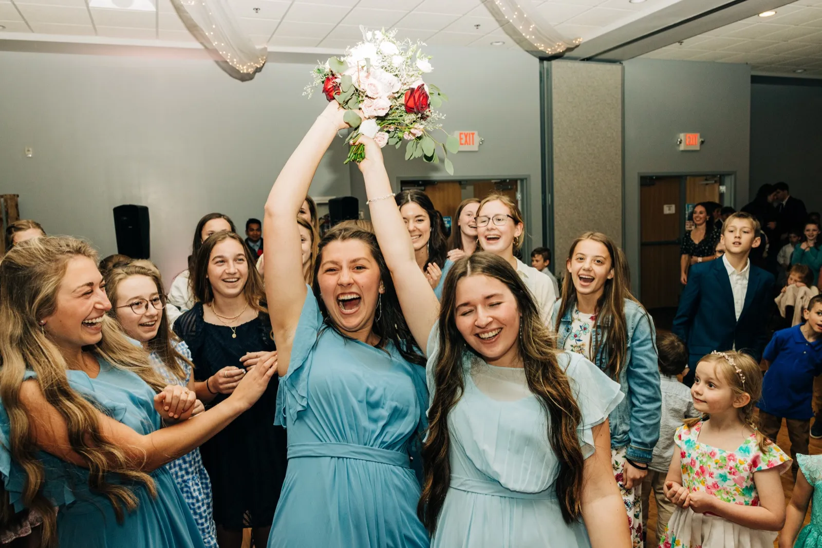 The catcher raises the bouquet overhead and shouts in triumph as a crowd of bridesmaids, kids, and guests laugh and cheer around her. — Tim Larsen Photography, Brainerd Lakes MN