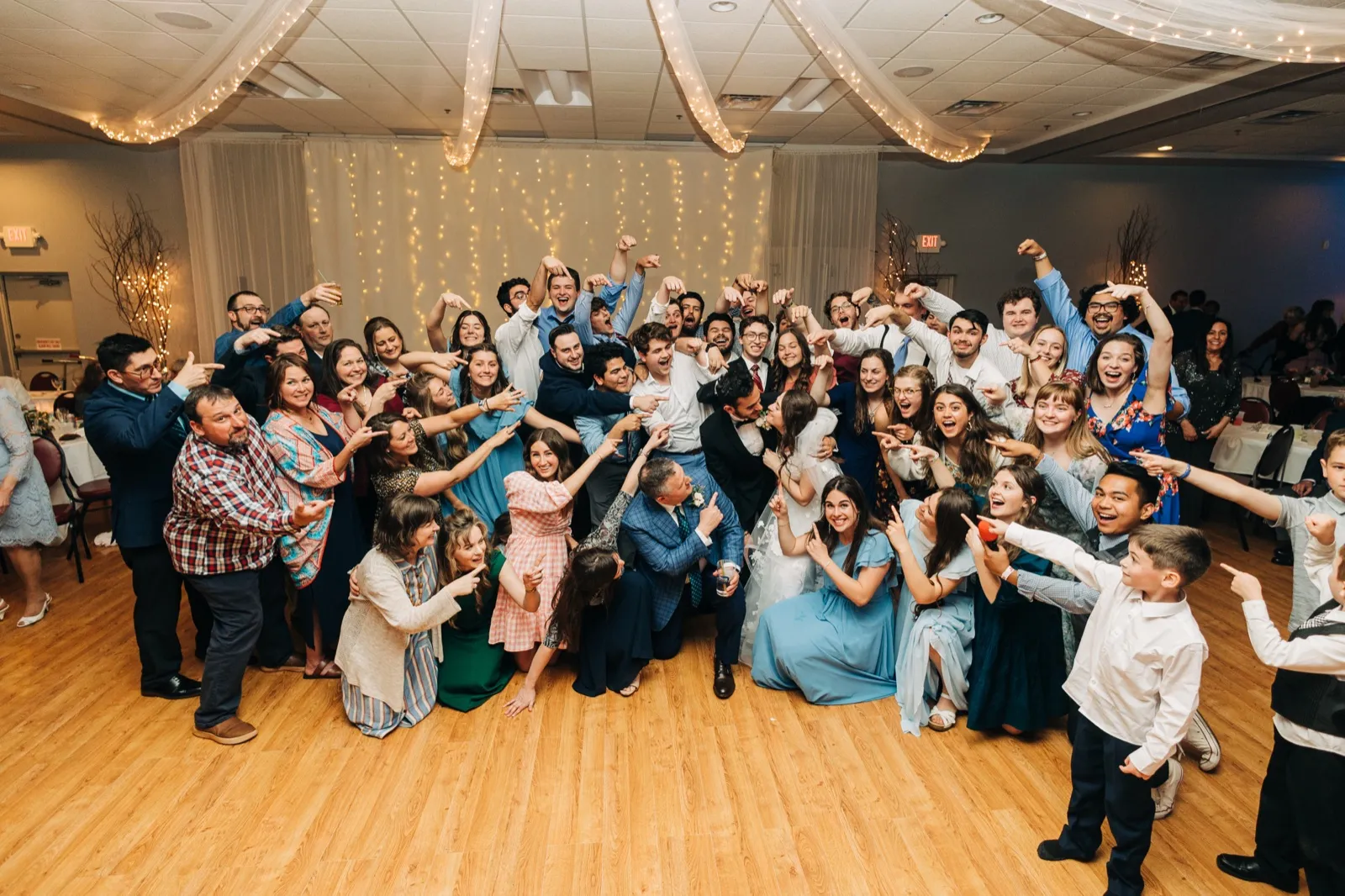 A large group of wedding guests crowds around the seated couple on a reception dance floor, all pointing toward them and cheering beneath draped fabric and string lights. — Tim Larsen Photography, Brainerd Lakes MN