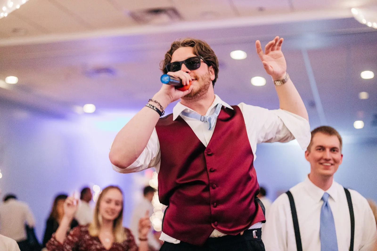 A guest in sunglasses and a maroon vest sings into a microphone with one hand raised on the reception dance floor, with other guests visible behind him. — Tim Larsen Photography, Brainerd Lakes MN