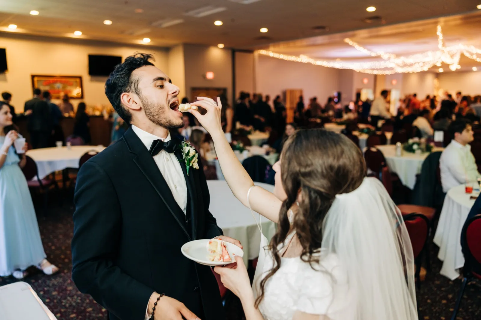 The bride reaches up to feed the groom a bite of cake during the reception, with string lights and guests at tables visible behind them. — Tim Larsen Photography, Brainerd Lakes MN