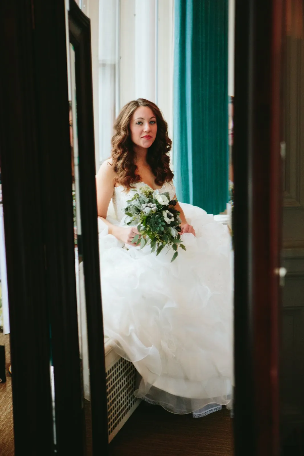 Framed through a doorway, the dressed bride seated at a window with bouquet in her lap and a teal curtain accent — Greysolon Plaza — Tim Larsen Photography, Duluth MN