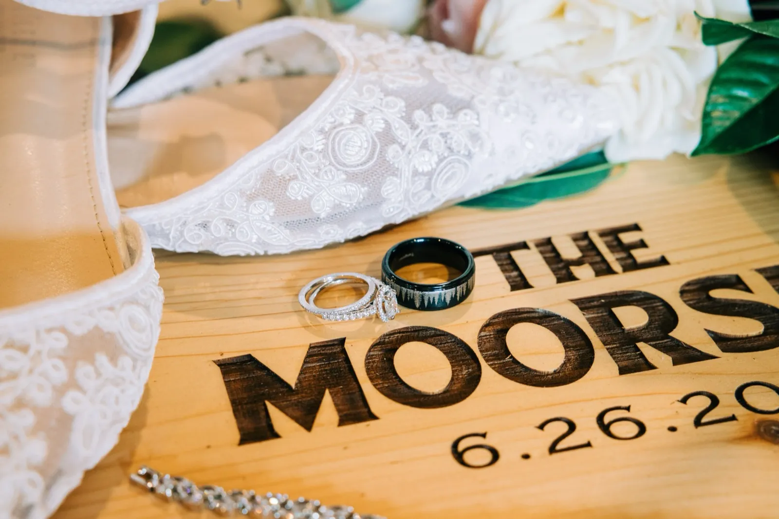 The couple's wedding rings resting on a hand-burned 'The Moorse' wood sign beside a lace bridal heel and a burgundy-and-blush bouquet — Tim Larsen Photography, Brainerd Lakes MN