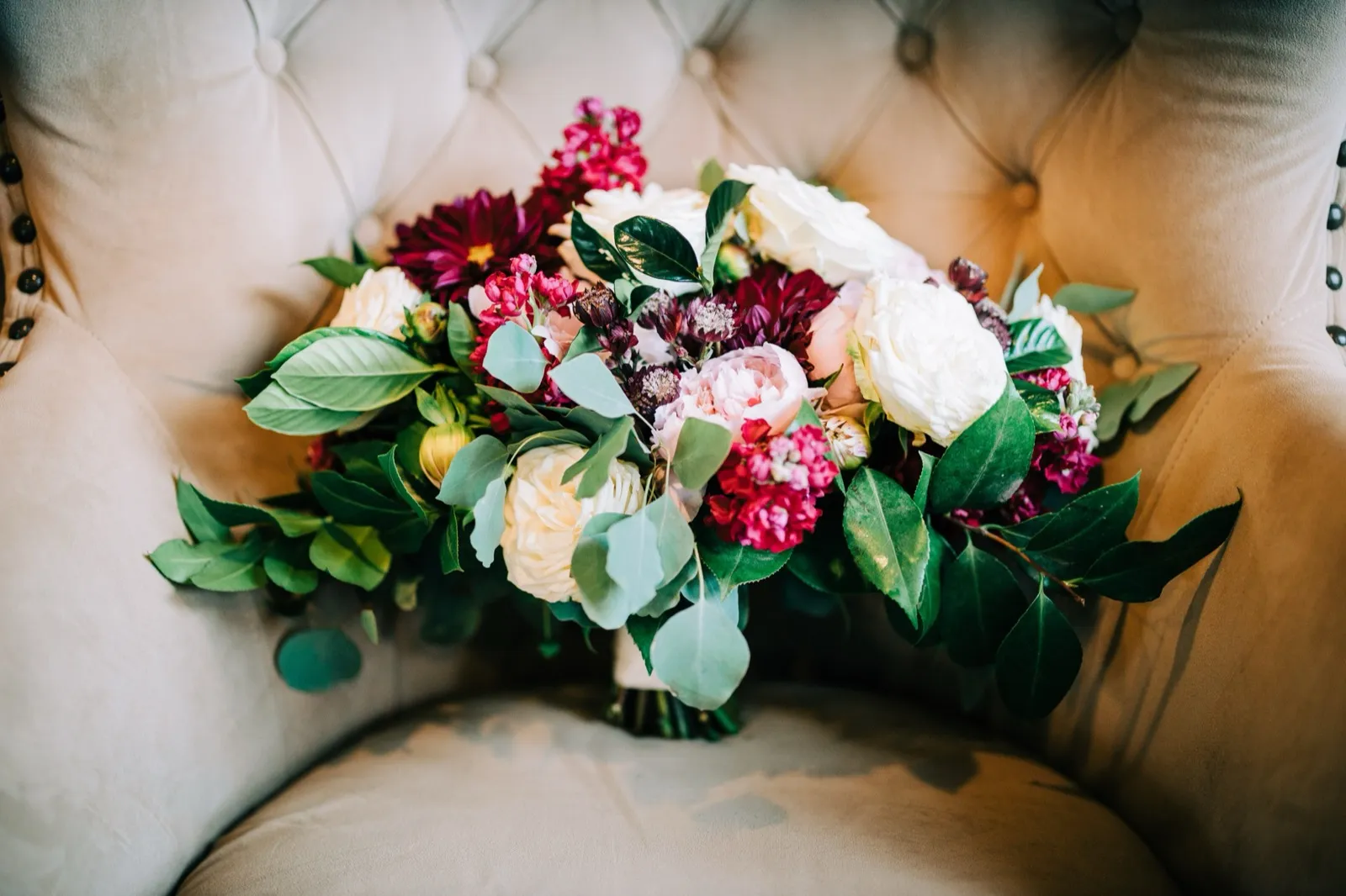 A bouquet of burgundy dahlias, blush garden roses, ivory peonies, and trailing eucalyptus resting on a cream velvet chair — Tim Larsen Photography, Brainerd Lakes MN