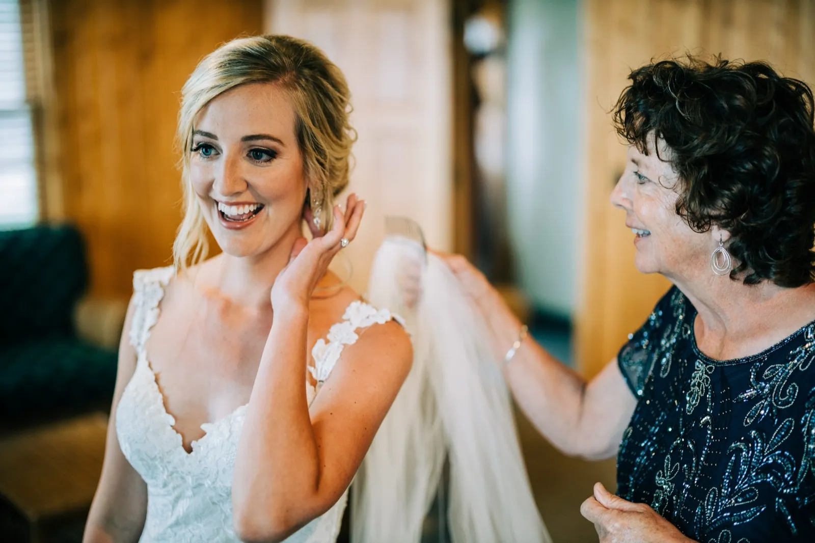 Shannon laughs while her mother, in a beaded navy dress, holds the veil ready beside her in a pine-paneled Madden's getting-ready room — Tim Larsen Photography, Brainerd Lakes MN