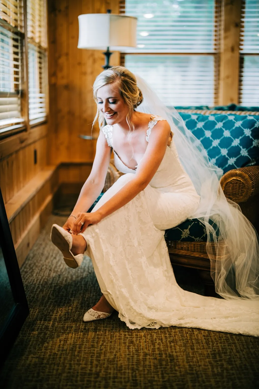 Shannon laughs as she leans down to fasten her shoe, her lace gown and veil spilling across a patterned settee in a wood-paneled cabin — Tim Larsen Photography, Brainerd Lakes MN