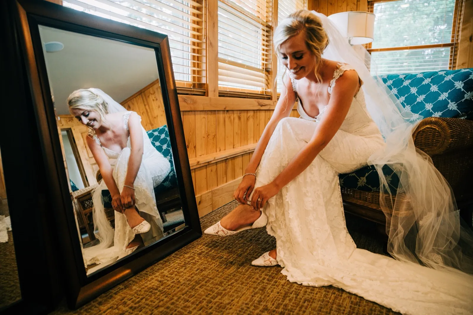 Shannon slips on a lace heel on a wicker sofa, her reflection mirrored in a tall standing mirror in the wood-paneled suite — Tim Larsen Photography, Brainerd Lakes MN