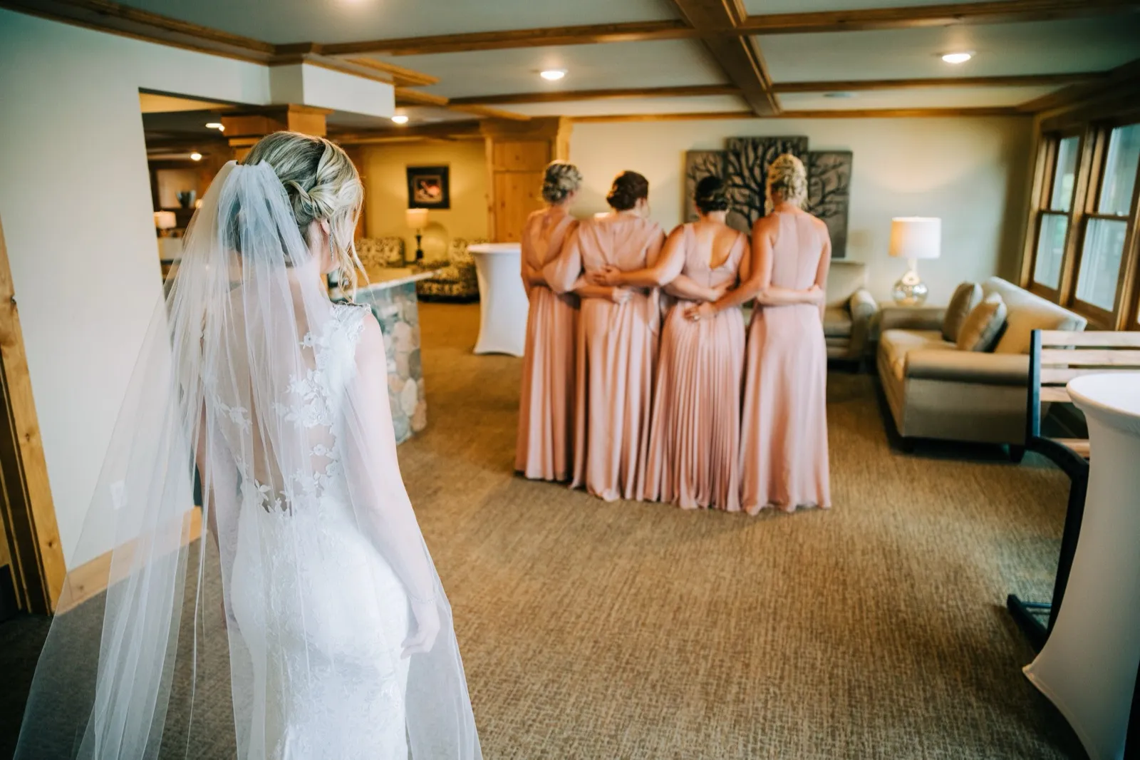 Shannon walks toward her bridesmaids, who stand with their backs turned in a timbered lodge lounge, waiting for the dress reveal — Tim Larsen Photography, Brainerd Lakes MN