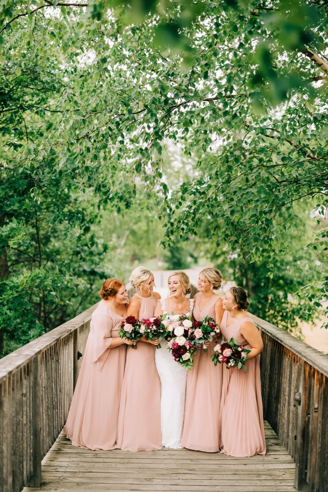 Shannon centered on a wooden footbridge holding her bouquet while four bridesmaids in blush laugh beneath a canopy of green leaves at Madden's — Tim Larsen Photography, Brainerd Lakes MN