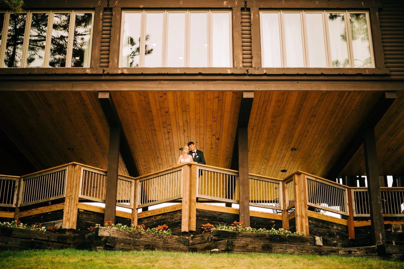 Shannon and Zach stand small at the center of a large timber-framed lodge balcony, the architecture rising symmetrically around them — Tim Larsen Photography, Brainerd Lakes MN