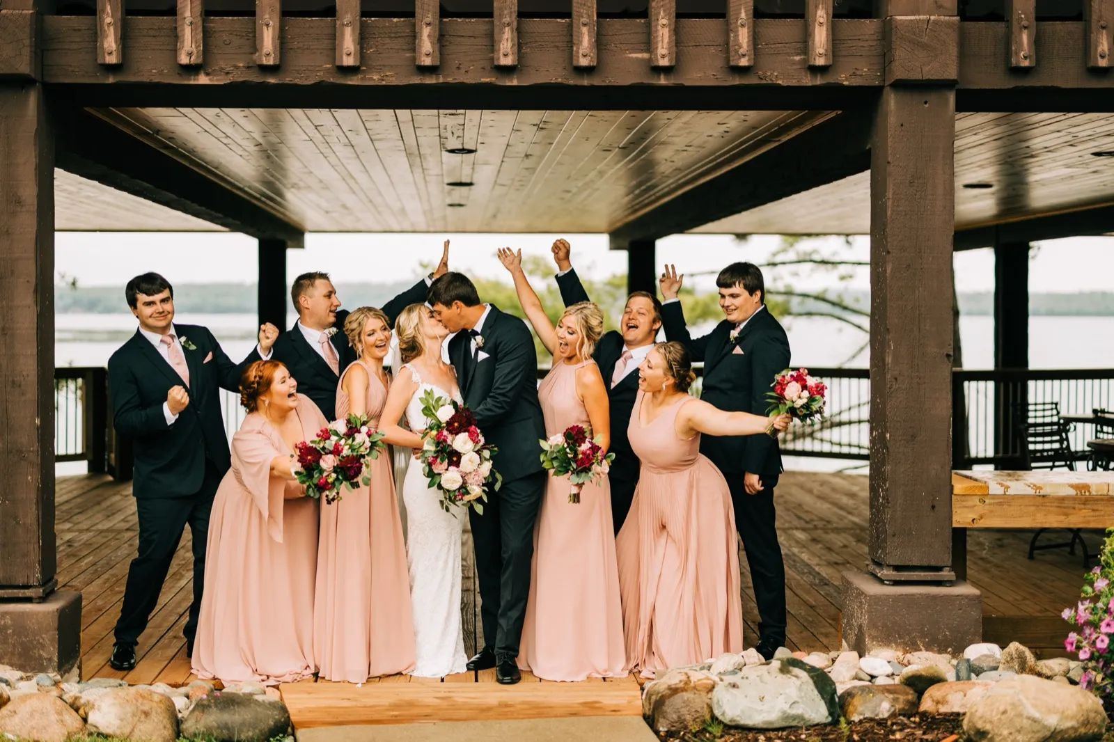 Shannon and Zach kiss as their bridal party cheers with raised arms beneath the timbered lakeside pavilion at Madden's on Gull Lake — Tim Larsen Photography, Brainerd Lakes MN