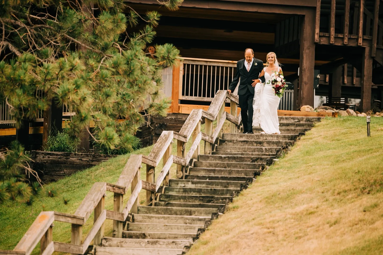 Shannon and her father descend the long lodge staircase toward the lakeside ceremony, a guest peeking from a window above — Tim Larsen Photography, Brainerd Lakes MN