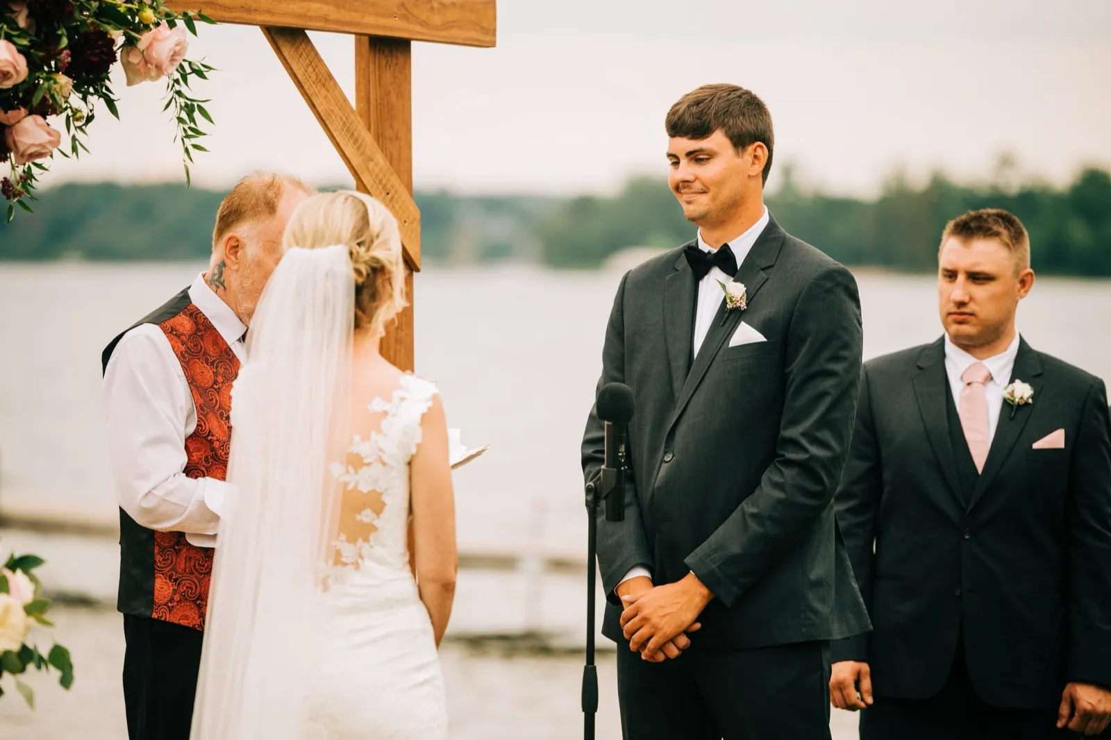 Zach looks at Shannon beneath a wooden ceremony arch with the officiant beside him and Gull Lake stretching out behind — Tim Larsen Photography, Brainerd Lakes MN